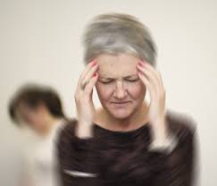 A woman with short grey hair stands with her eyes closed, pressing her hands to her temples as if experiencing dizziness or a headache. The background is blurred, emphasizing her discomfort and unsteady feeling.