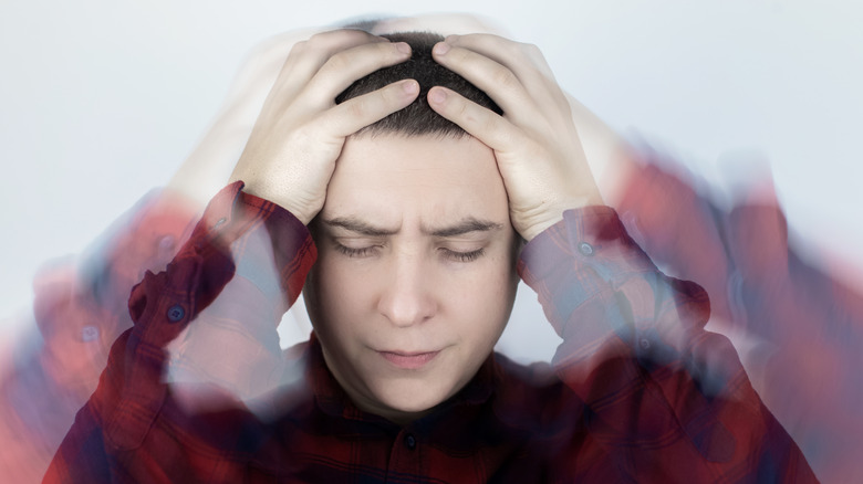 A person with short dark hair holds their head with both hands, eyes closed in discomfort, as blurred motion surrounds them, creating a visual effect that suggests dizziness or vertigo.